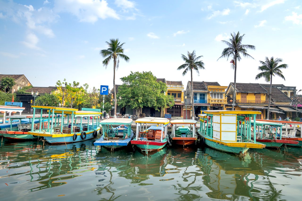 Colorful excursion boats moored on river near embankment near palm trees and typical houses on sunny day in Hoi An